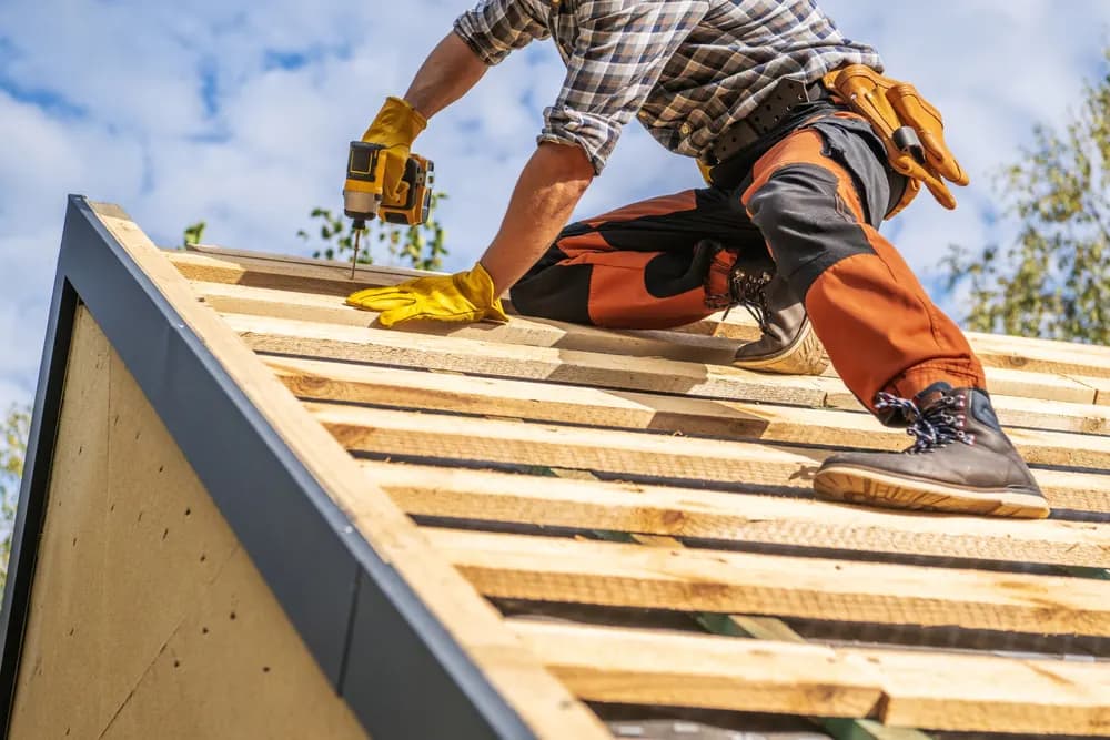 Construction worker in high-visibility clothing using a power drill to secure wooden battens on a roof frame under a blue sky